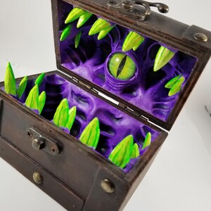 Puede incluir: Un cofre del tesoro de madera con tapa con bisagras y herrajes met&aacute;licos. El interior revela un dise&ntilde;o morado texturizado con un gran ojo verde y dientes verdes puntiagudos. El cofre abierto se asemeja a la boca de un monstruo.