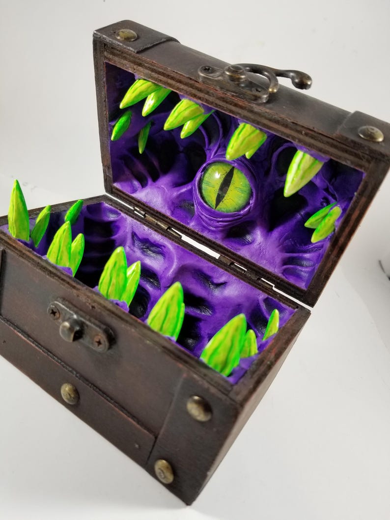 Puede incluir: Un cofre del tesoro de madera con tapa con bisagras y herrajes met&aacute;licos. El interior revela un dise&ntilde;o morado texturizado con un gran ojo verde y dientes verdes puntiagudos. El cofre abierto se asemeja a la boca de un monstruo.