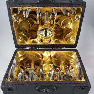 Puede incluir: Un cofre del tesoro de madera negra con tapa abisagrada, abierto para revelar un interior dorado con un dise&ntilde;o de monstruo. Dientes plateados bordean los bordes, y gemas coloridas y una espada son visibles en el interior. Un cierre met&aacute;lico asegura la tapa.