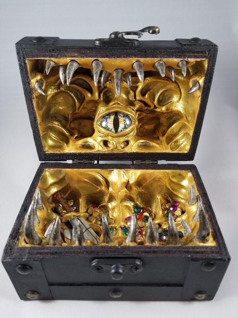Puede incluir: Un cofre del tesoro de madera negra con tapa abisagrada, abierto para revelar un interior dorado con un dise&ntilde;o de monstruo. Dientes plateados bordean los bordes, y gemas coloridas y una espada son visibles en el interior. Un cierre met&aacute;lico asegura la tapa.