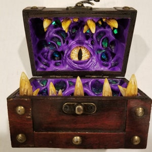 Puede incluir: Un cofre del tesoro de madera con tapa con bisagras, con un dise&ntilde;o amenazante. El interior es de color p&uacute;rpura vibrante con un ojo amarillo grande y dientes afilados amarillos. El cofre est&aacute; detallado con detalles met&aacute;licos.