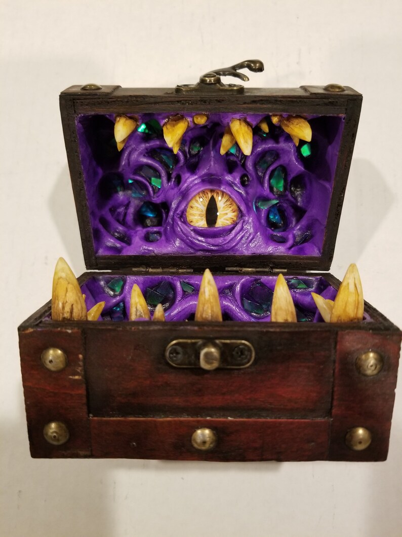 Puede incluir: Un cofre del tesoro de madera con tapa con bisagras, con un dise&ntilde;o amenazante. El interior es de color p&uacute;rpura vibrante con un ojo amarillo grande y dientes afilados amarillos. El cofre est&aacute; detallado con detalles met&aacute;licos.
