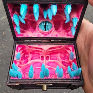 Puede incluir: Un peque&ntilde;o cofre de madera abierto, que revela un interior tem&aacute;tico de fantas&iacute;a. El interior rosa presenta dientes azules puntiagudos y un ojo azul central. El cofre est&aacute; abierto, mostrando el dise&ntilde;o &uacute;nico.