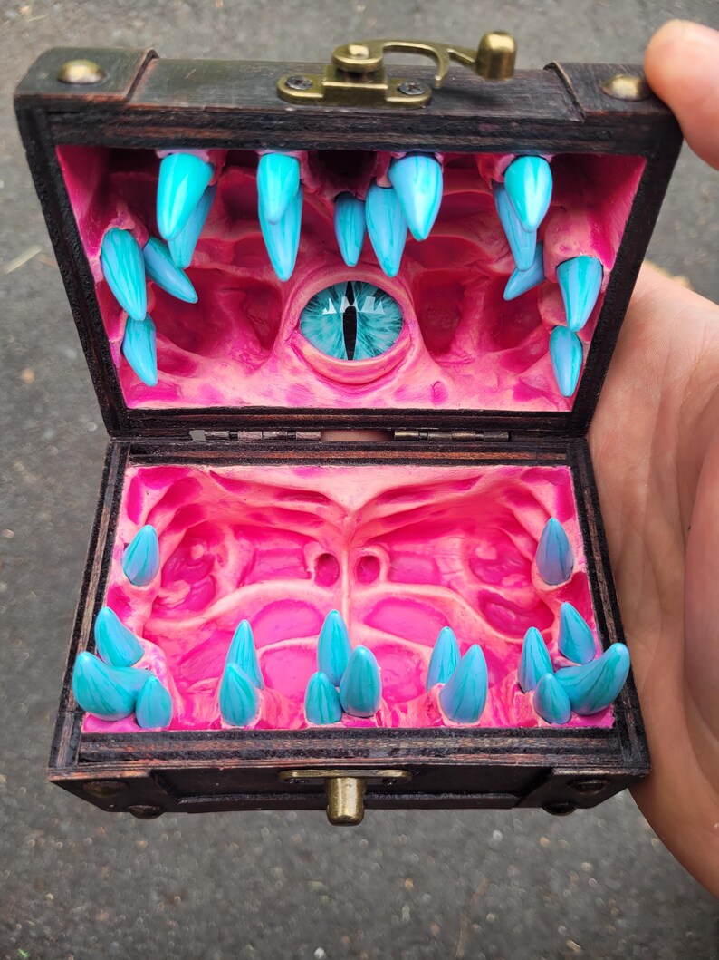 Puede incluir: Un peque&ntilde;o cofre de madera abierto, que revela un interior tem&aacute;tico de fantas&iacute;a. El interior rosa presenta dientes azules puntiagudos y un ojo azul central. El cofre est&aacute; abierto, mostrando el dise&ntilde;o &uacute;nico.