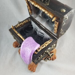 Puede incluir: Un cofre del tesoro decorativo con exterior de madera marr&oacute;n oscuro, detalles met&aacute;licos ornamentados y tapa con bisagras. El cofre est&aacute; abierto, revelando un interior negro y un objeto morado en forma de lengua. El cofre descansa sobre dos pies marrones.