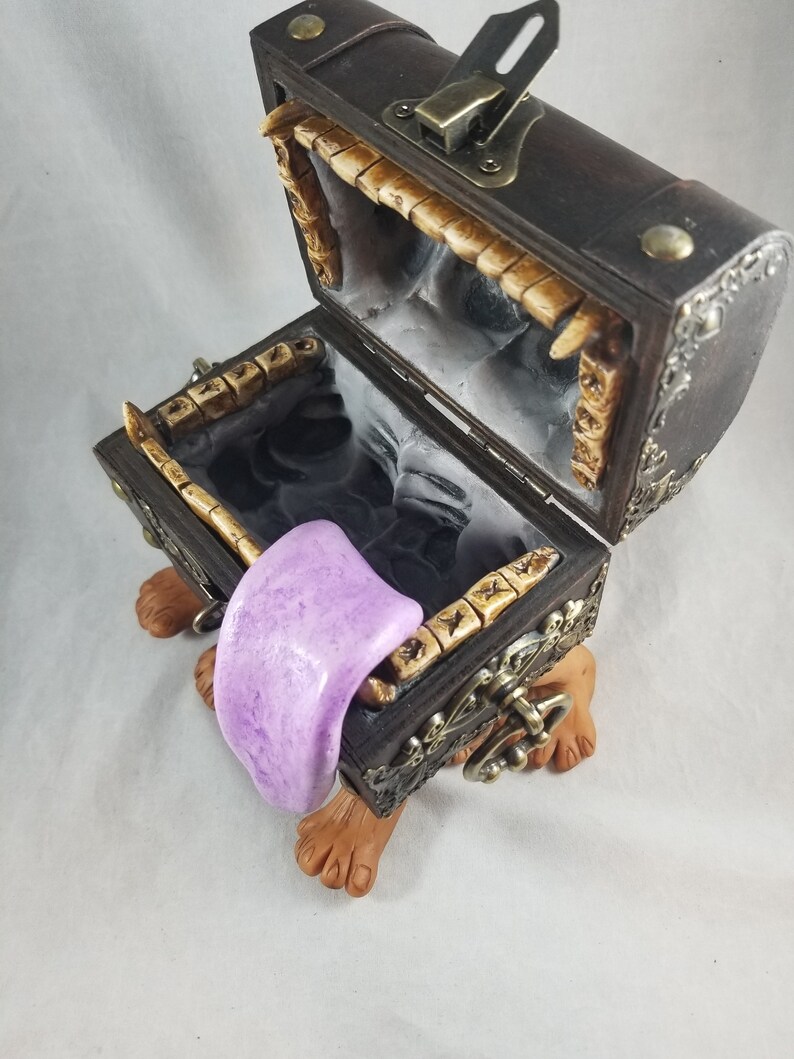 Puede incluir: Un cofre del tesoro decorativo con exterior de madera marr&oacute;n oscuro, detalles met&aacute;licos ornamentados y tapa con bisagras. El cofre est&aacute; abierto, revelando un interior negro y un objeto morado en forma de lengua. El cofre descansa sobre dos pies marrones.