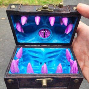 Puede incluir: Un cofre del tesoro de madera con tapa con bisagras, abierto para revelar un interior azul con un dise&ntilde;o de monstruo. El interior presenta colmillos morados y un ojo central. El cofre es marr&oacute;n con herrajes met&aacute;licos.