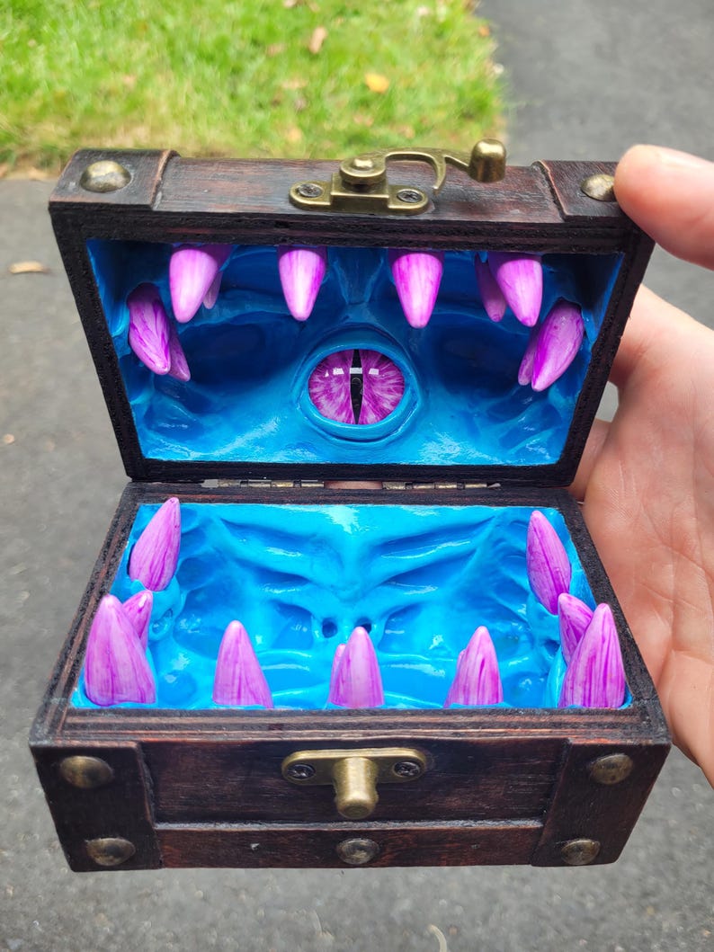 Puede incluir: Un cofre del tesoro de madera con tapa con bisagras, abierto para revelar un interior azul con un dise&ntilde;o de monstruo. El interior presenta colmillos morados y un ojo central. El cofre es marr&oacute;n con herrajes met&aacute;licos.