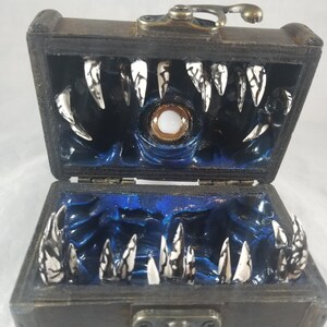 Puede incluir: Un cofre del tesoro de madera, abierto para revelar un interior azul oscuro con un ojo central y filas de dientes afilados blancos y negros. El cofre tiene una tapa con bisagras y herrajes met&aacute;licos desgastados.