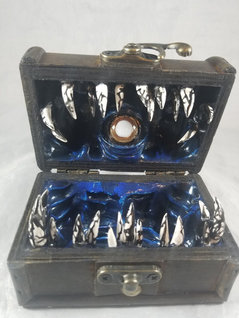 Puede incluir: Un cofre del tesoro de madera, abierto para revelar un interior azul oscuro con un ojo central y filas de dientes afilados blancos y negros. El cofre tiene una tapa con bisagras y herrajes met&aacute;licos desgastados.