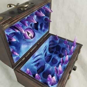 Puede incluir: Un cofre del tesoro de madera con un exterior marr&oacute;n oscuro y una tapa con bisagras. El interior est&aacute; pintado de azul y presenta una cara de monstruo esculpida con un ojo morado y dientes afilados en forma de cristal morado.