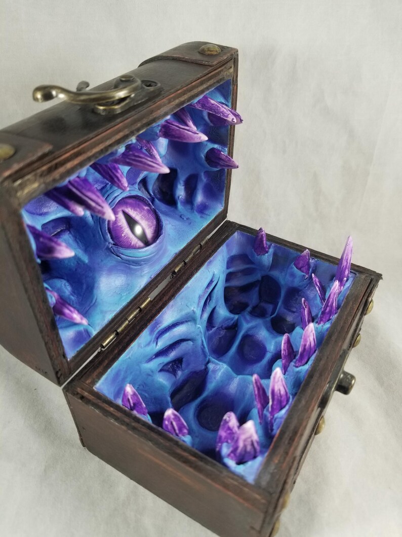 Puede incluir: Un cofre del tesoro de madera con un exterior marr&oacute;n oscuro y una tapa con bisagras. El interior est&aacute; pintado de azul y presenta una cara de monstruo esculpida con un ojo morado y dientes afilados en forma de cristal morado.