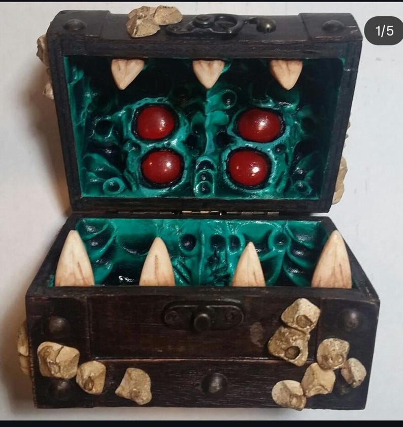 Puede incluir: Un cofre del tesoro de madera con exterior marr&oacute;n oscuro e interior turquesa. El interior presenta grandes formas redondas rojas y dientes blancos y puntiagudos. El cofre est&aacute; decorado con peque&ntilde;as piedras de color beige.