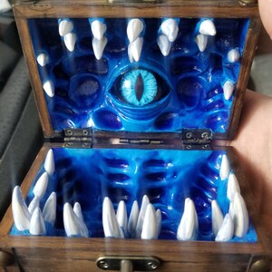 Puede incluir: Cofre del tesoro de madera abierto, con un interior azul con un gran ojo y filas de dientes blancos. El cofre es de color marr&oacute;n oscuro con herrajes met&aacute;licos y un cierre.