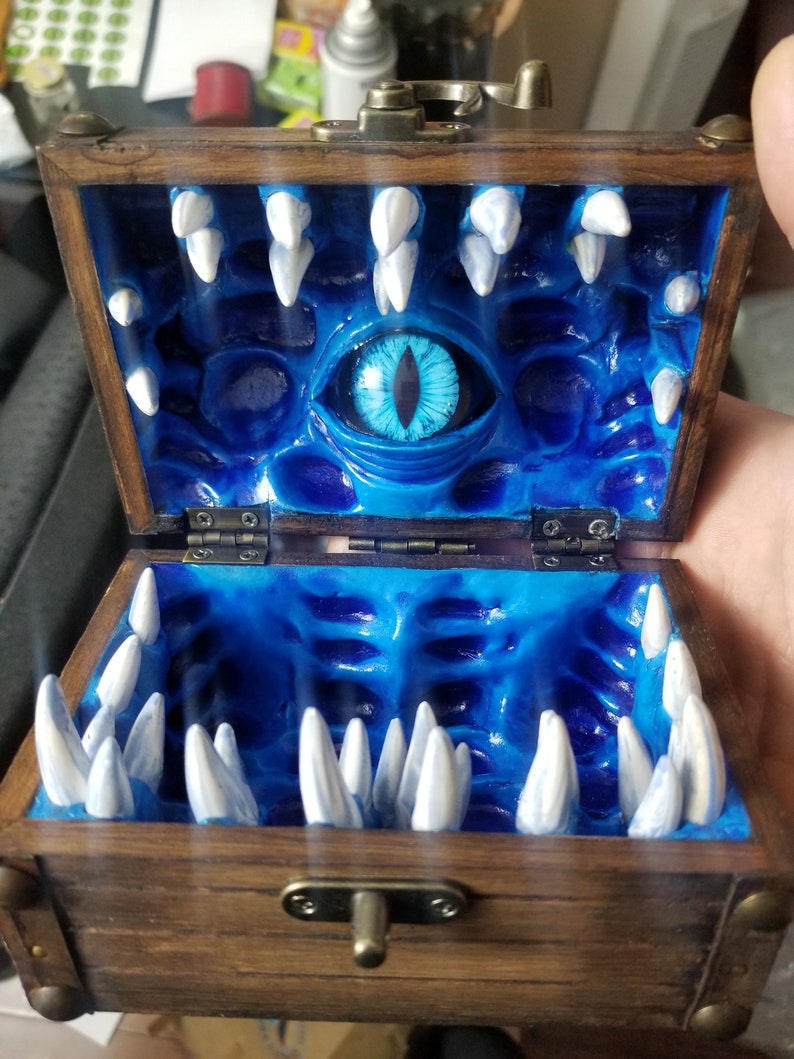 Puede incluir: Cofre del tesoro de madera abierto, con un interior azul con un gran ojo y filas de dientes blancos. El cofre es de color marr&oacute;n oscuro con herrajes met&aacute;licos y un cierre.