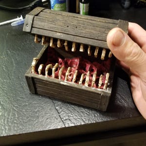 Puede incluir: Un cofre del tesoro de madera con tapa con bisagras, detallado con un dise&ntilde;o de mand&iacute;bula esquel&eacute;tica. El interior est&aacute; forrado con tela roja, y el cofre est&aacute; abierto, revelando los dientes esquel&eacute;ticos. El cofre es sostenido por una mano.