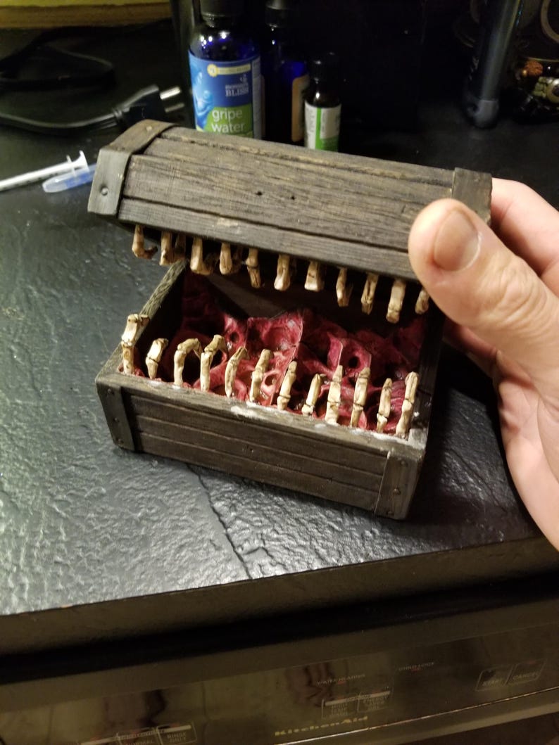 Puede incluir: Un cofre del tesoro de madera con tapa con bisagras, detallado con un dise&ntilde;o de mand&iacute;bula esquel&eacute;tica. El interior est&aacute; forrado con tela roja, y el cofre est&aacute; abierto, revelando los dientes esquel&eacute;ticos. El cofre es sostenido por una mano.