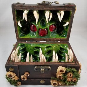 Puede incluir: Un cofre de madera marr&oacute;n oscuro con tapa abierta, que revela un interior verde con grandes dientes blancos y tres gemas rojas. El exterior est&aacute; decorado con musgo y peque&ntilde;os detalles en forma de hongo.