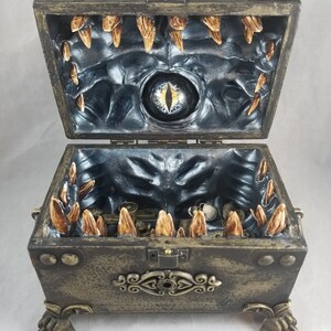 Puede incluir: Un cofre del tesoro abierto de estilo antiguo con un interior tem&aacute;tico de monstruos. El cofre es de color bronce con detalles decorativos y patas en forma de garra. En el interior, un dise&ntilde;o oscuro y texturizado presenta un ojo central y estructuras en forma de dientes.