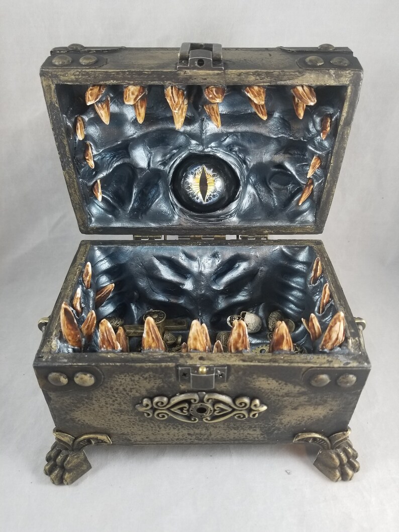 Puede incluir: Un cofre del tesoro abierto de estilo antiguo con un interior tem&aacute;tico de monstruos. El cofre es de color bronce con detalles decorativos y patas en forma de garra. En el interior, un dise&ntilde;o oscuro y texturizado presenta un ojo central y estructuras en forma de dientes.