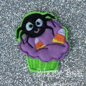 Puede incluir: Un cupcake morado con glaseado verde y maíz dulce en la parte superior. Una araña negra asoma desde el cupcake.