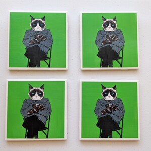 May include: Set of four square coasters featuring a grumpy cat wearing a blue jacket and holding a pair of mittens. The coasters have a green background.