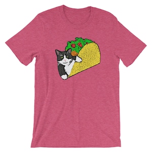 May include: A pink t-shirt with a black and white cat inside a yellow taco. The cat is looking at the viewer with a mischievous expression. The taco is filled with green lettuce and a red tomato.