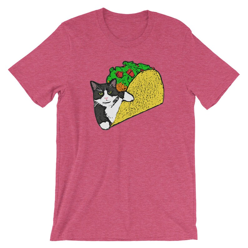 May include: A pink t-shirt with a black and white cat inside a yellow taco. The cat is looking at the viewer with a mischievous expression. The taco is filled with green lettuce and a red tomato.
