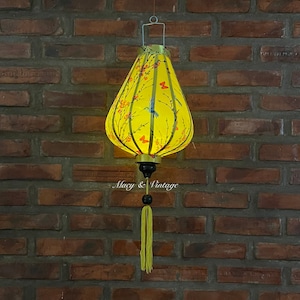 Puede incluir: Una linterna amarilla iluminada con un diseño floral y de mariposas. La linterna está colgada contra una pared de ladrillos. La linterna tiene una base negra y dorada con una borla amarilla.