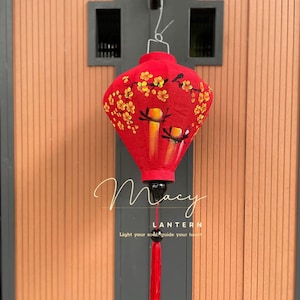 May include: Red Chinese lantern with a floral design and a tassel. The lantern is hanging from a hook on a wooden door. The text "Macy Lantern Light your soul, guide your heart" is visible below the lantern.