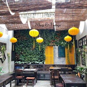 May include: A patio dining area with wooden tables and chairs, surrounded by lush greenery and yellow lanterns hanging from a bamboo roof. The sign "Macy & Vintage" is visible above the greenery.