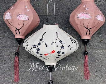 Puede incluir: Tres linternas de seda pintadas a mano con borlas. Las linternas tienen forma de diamante y tienen un fondo blanco con diseños florales negros y rojos. Las linternas están colgadas del techo.
