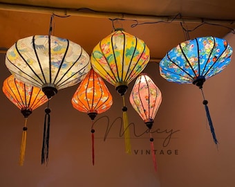 May include: A collection of colorful, illuminated paper lanterns hanging from a ceiling. The lanterns feature floral and nature-inspired designs in shades of orange, yellow, pink, and blue. Each lantern has a tassel hanging from the bottom.