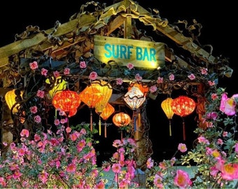 May include: A wooden structure with a sign reading SURF BAR, decorated with intertwined branches and pink flowers. Multiple glowing paper lanterns in red, orange, and yellow hang from the frame, illuminating the dark night setting.