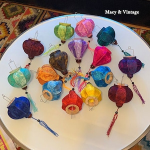 Pode incluir: Uma coleção de 15 lanternas de tecido coloridas com borlas. As lanternas são de várias formas e tamanhos, incluindo redondas, ovais e em forma de lágrima. As lanternas são feitas de uma variedade de tecidos, incluindo seda, veludo e algodão. As lanternas estão penduradas em um teto branco.