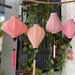 May include: Four hanging lanterns in varying shades of pink, each featuring a fabric-covered frame, a black base, and a long matching pink tassel. The lanterns are suspended from a horizontal wooden branch, showcasing different shapes and sizes.