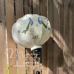 May include: A white silk lantern with a black metal frame and a tassel. The lantern is decorated with a painted design of two peacocks and green leaves. The text "Macy Lantern" and "Light your soul, guide your heart" is printed on the lantern.