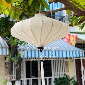 Pode incluir: Uma lanterna creme com um padrão floral pendurada num galho de árvore. A lanterna tem uma base preta e é suspensa por um fino cordão branco. O texto "Macy Lantern" é visível na imagem, juntamente com a frase "Light your soul, guide your heart."