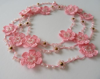 May include: A long necklace featuring light pink crocheted flowers interspersed with small, translucent pink beads and metallic gold-toned accents. The delicate floral elements are connected by a thin, matching pink thread in a continuous strand.