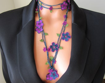 以下が含まれることがあります： 手作りのフローラルネックレス。紫とティールの鉤針編みの花、緑の葉、ピンクのビーズクラスターが特徴です。黒いブレザーの上から着用されています。