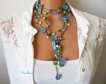 May include: A handmade crochet necklace featuring blue flower accents, white stone beads, and green vines. The necklace is worn over a white top and is a unique piece of jewelry.