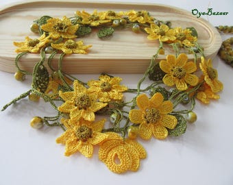 May include: A handmade yellow crochet necklace featuring daisy-like flowers and green leaves. The necklace is adorned with small, round beads and is displayed on a wooden tray. The Oya Bazaar logo is visible in the top right corner.