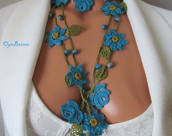 Può includere: Una lunga collana all'uncinetto con fiori blu dal centro giallo, foglie verdi lavorate all'uncinetto e piccole perline blu su un sottile cordino verde. Il testo Oya Bazaar è stampato in blu sul lato sinistro dell'immagine.