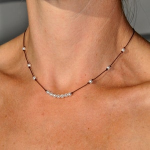 May include: Delicate pearl necklace featuring small, round, white pearls strung on a dark brown cord. The necklace has a simple design with pearls evenly spaced along the cord, creating a minimalist and elegant look. The necklace is shown around a person's neck.