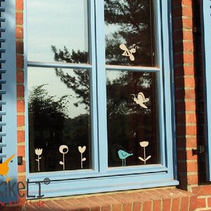 May include: A blue window with white silhouettes of flowers, birds, and a bee on the glass. The window is framed in blue wood and is set in a brick wall. The text "KikeLi" is visible in the bottom left corner of the image.