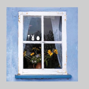 May include: A white window with a blue frame, featuring a flower, bunny, and egg Easter decorations on the glass.  Yellow flowers are visible through the window.