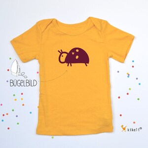 Könnte beinhalten: Ein gelbes T-Shirt mit kurzen Ärmeln und einem burgunderfarbenen Bügelbild einer Marienkäfer. Der Text "Bügelbild" ist links im Bild zu sehen.