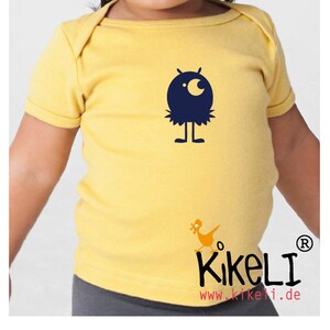 May include: A yellow short-sleeved t-shirt with a blue monster graphic and the text "KikeLI www.kikeli.de" on the bottom.