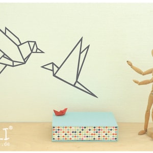May include: Two origami paper birds in flight, one in front of the other, against a white wall. A small red origami boat sits on a blue box with a colorful patterned bottom. A wooden artist's mannequin stands to the right of the box, with its arms outstretched.