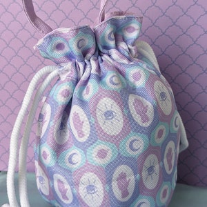 Puede incluir: Una pequeña bolsa con cordón con un patrón repetido de círculos blancos, azules y morados. La bolsa tiene un asa morada a juego y un cierre de cordón blanco. El fondo es de color morado claro.